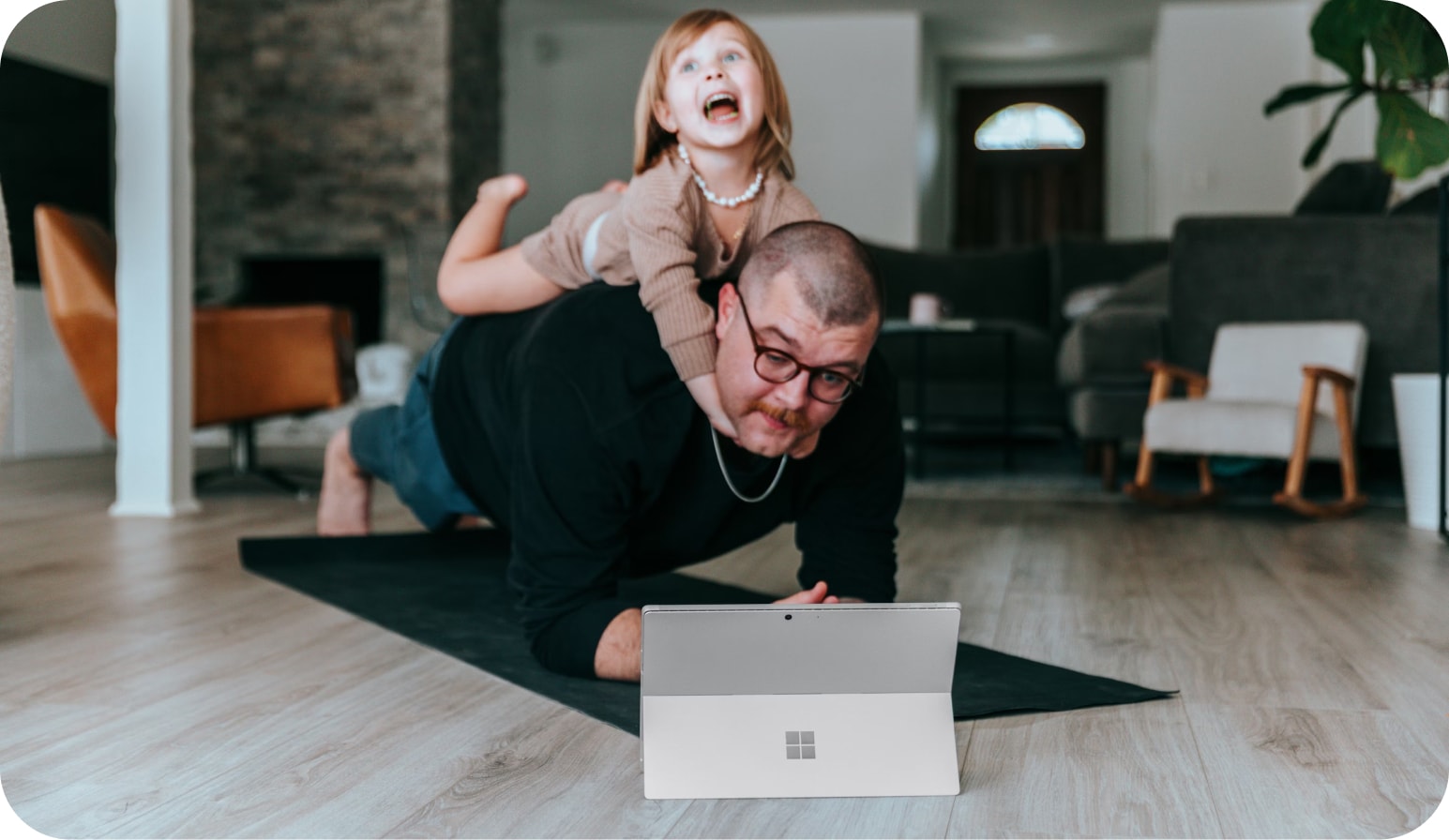 En mann ligger på en yogamatte i et hjemlig miljø, mens et barn sitter på ryggen hans med et stort smil. Foran mannen er det en bærbar datamaskin, og de ser ut til å være engasjert i en aktivitet sammen. Rommet er innredet med moderne møbler, inkludert en stol og en sofa