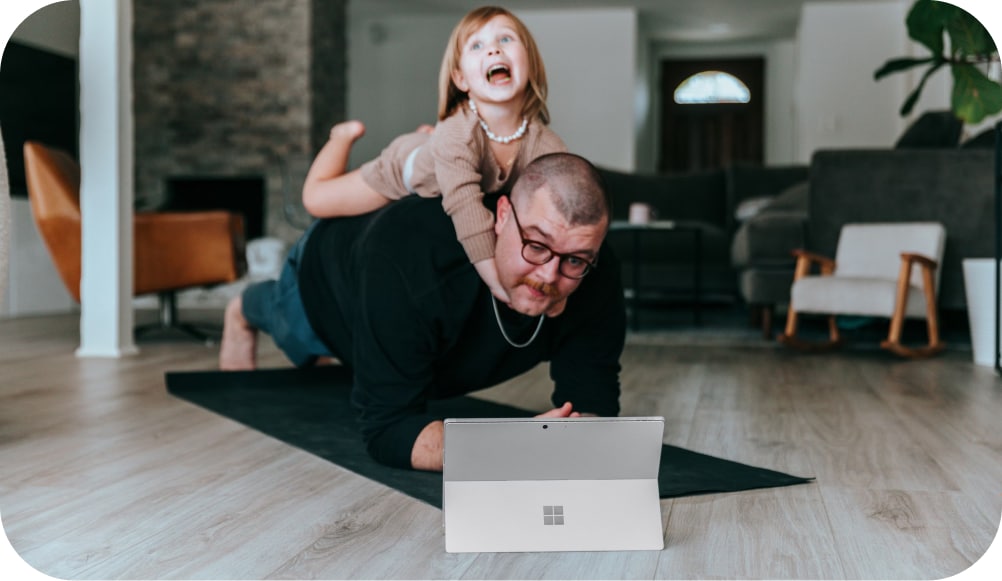 En mann ligger på en yogamatte i et hjemlig miljø, mens et barn sitter på ryggen hans med et stort smil. Foran mannen er det en bærbar datamaskin, og de ser ut til å være engasjert i en aktivitet sammen. Rommet er innredet med moderne møbler, inkludert en stol og en sofa